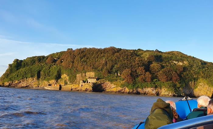 Steep Holm Island surrounded by water. People in boat looking at island which is steep and covered in trees and foliage. Remains of buildings can be s